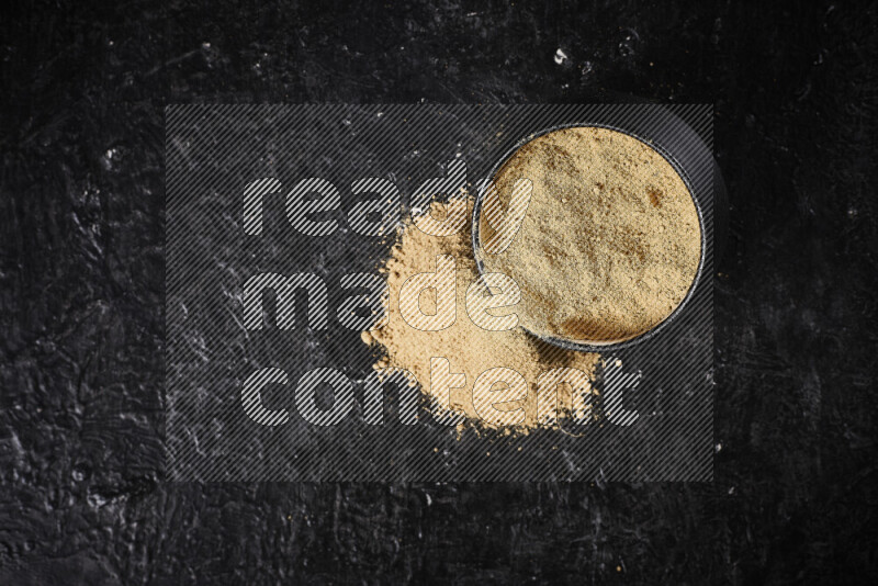 A black pottery bowl full of ground ginger powder with fallen powder from it on black background