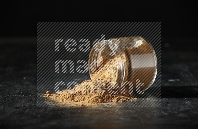 A flipped glass jar full of allspice powder and powder spilled out of it on a textured black flooring