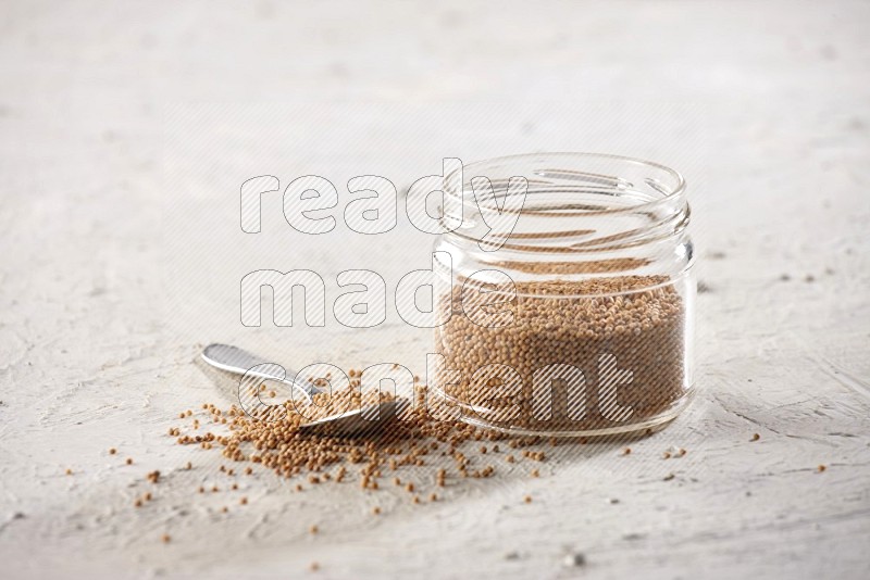A glass jar and a metal spoon full of mustard seeds on a textured white flooring