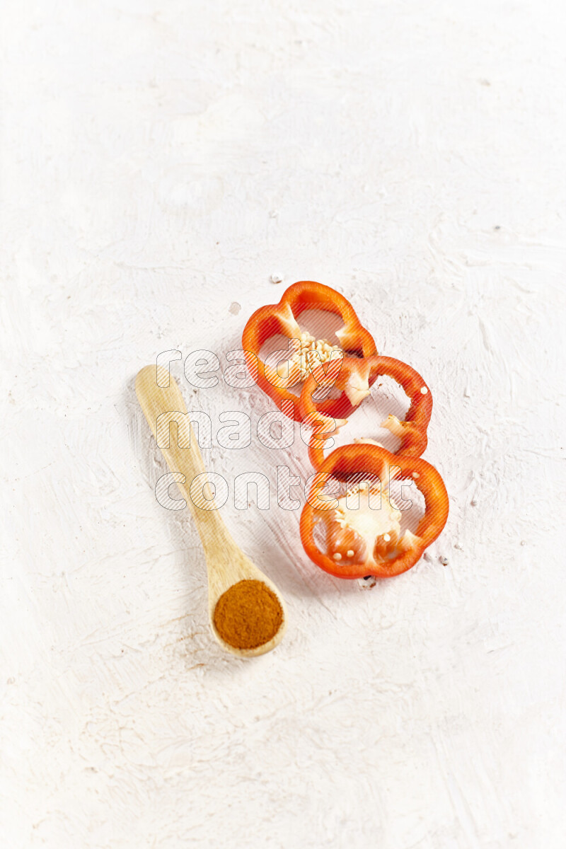 A wooden spoon full of ground paprika powder with red bell pepper slices beside it on white background