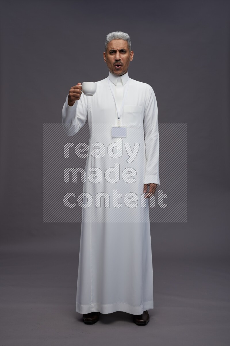 Saudi man wearing thob with neck strap employee badge standing holding mug on gray background