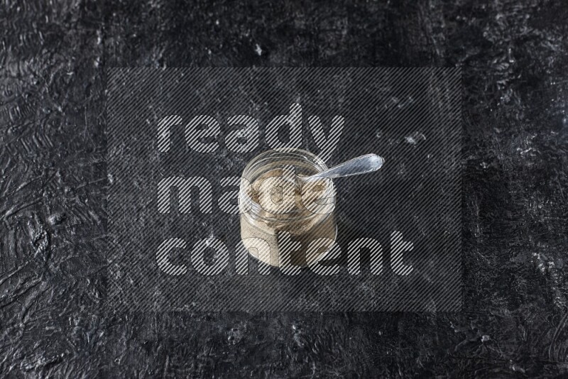 A glass jar and a metal spoon full of white pepper powder on textured black flooring