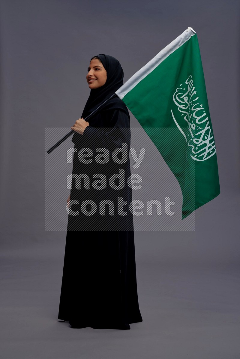 Saudi woman wearing Abaya standing holding Saudi flag on gray background