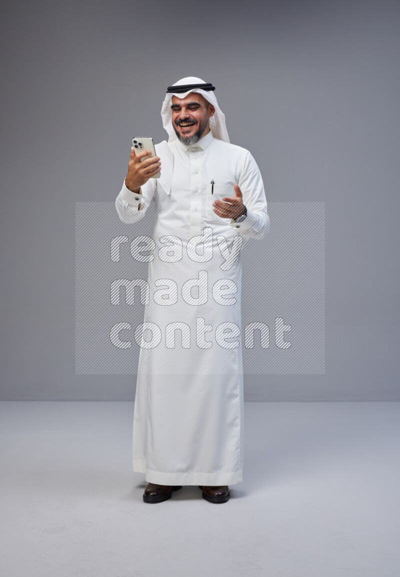 Saudi man Wearing Thob and white Shomag standing texting on phone on Gray background
