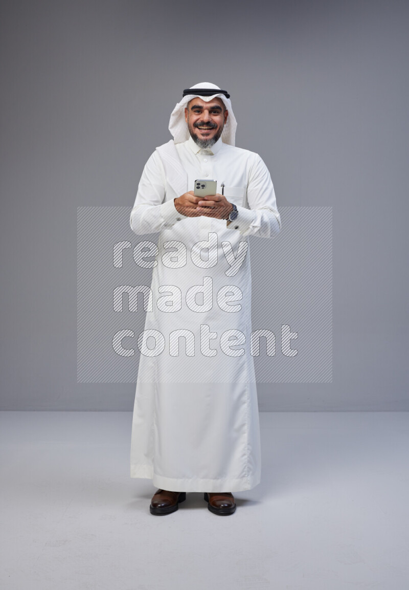 Saudi man Wearing Thob and white Shomag standing texting on phone on Gray background