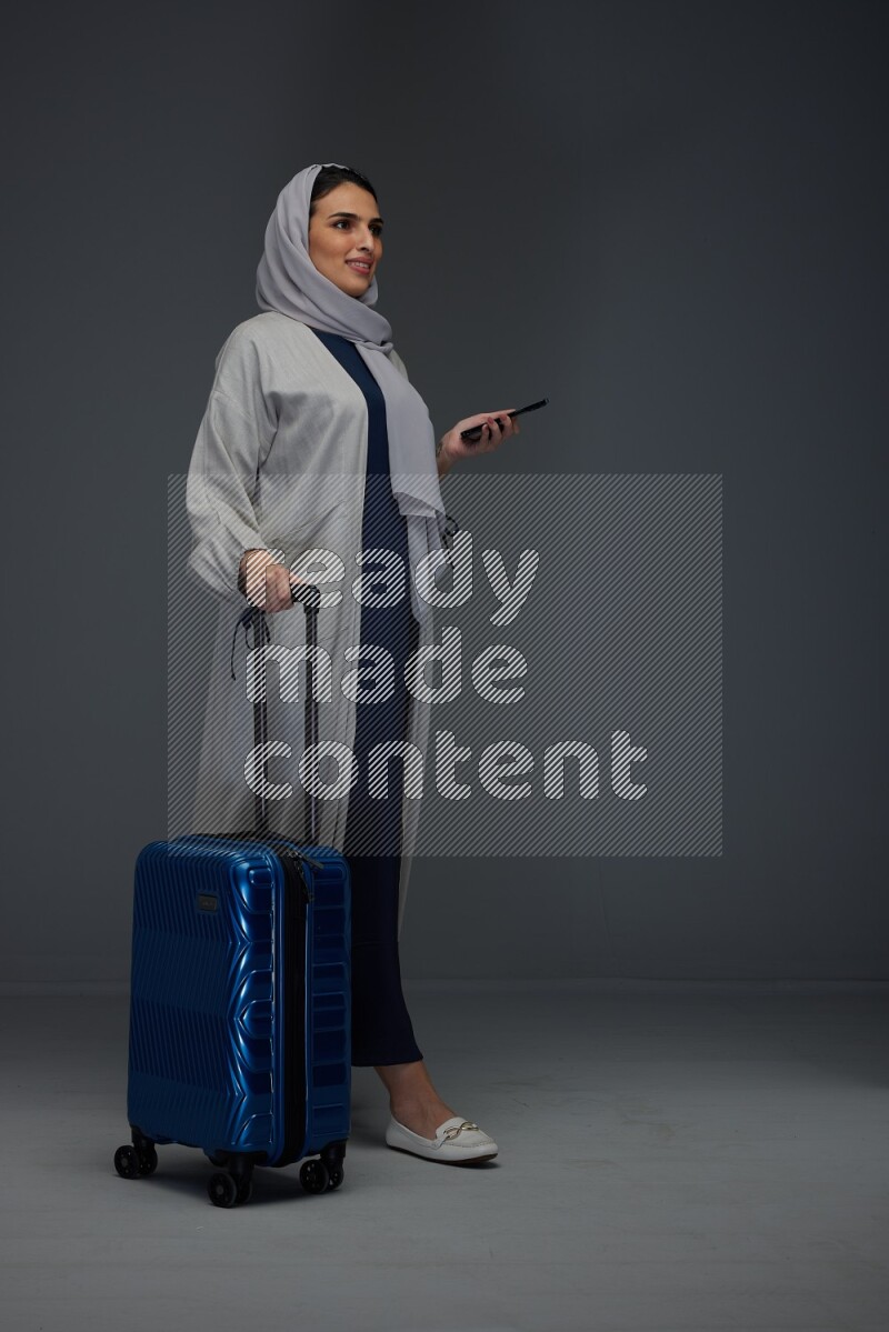 A Saudi woman wearing a light gray Abaya and head scarf standing holding a phone and being shocked  eye level on a grey background