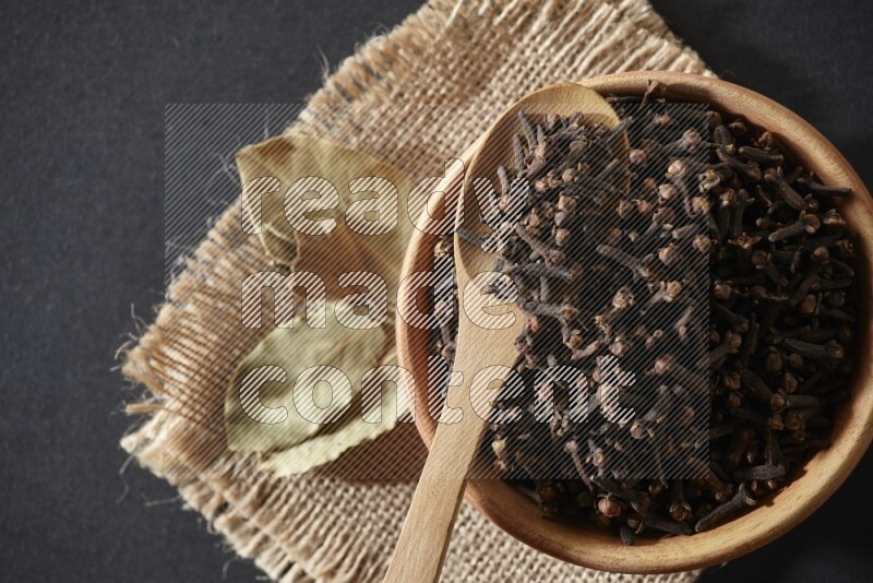 A wooden bowl, a wooden spoon full of cloves, and bay leaves (laurel) on a piece of burlap on a black flooring