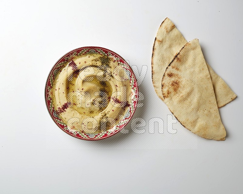 Hummus in a red plate with patterns garnished with zaatar & sumak on a white background