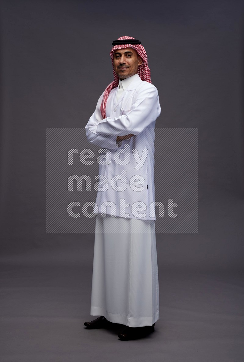 Saudi man wearing thob with lab coat and shomag with pocket employee badge standing with crossed arms on gray background