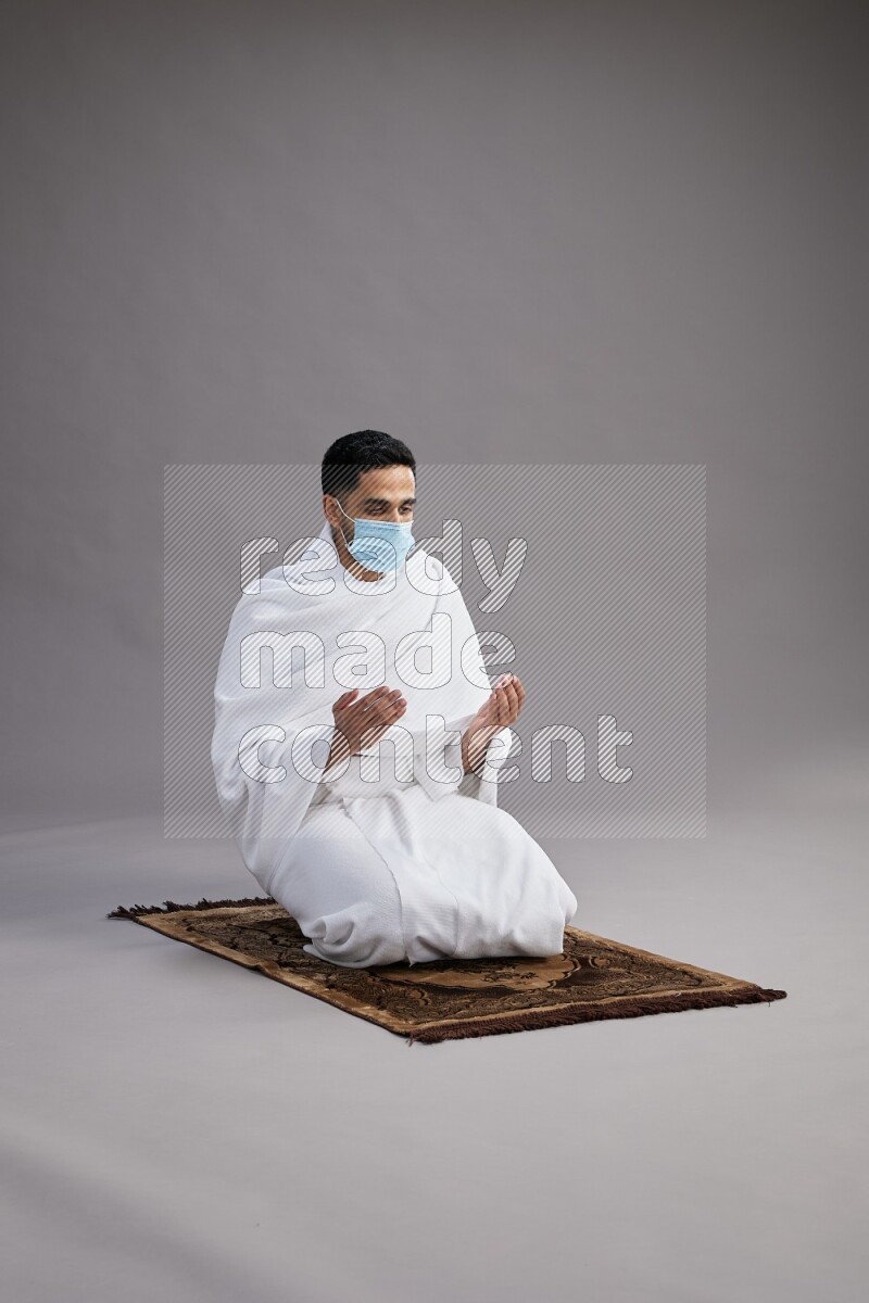 A man wearing Ehram with face mask sitting on floor performing dua'a on gray background