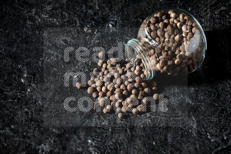 A flipped glass spice jar full of allspice whole balls and the balls spilled out of it on a textured black flooring
