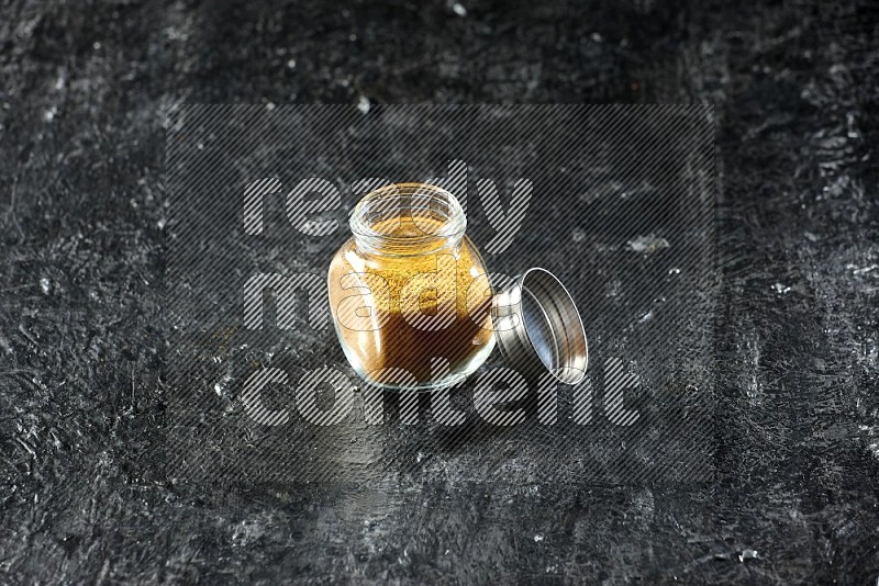 A glass spice jar full of turmeric powder on a textured black flooring