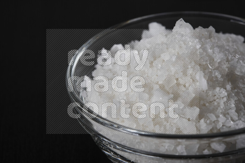 A glass bowl full of coarse sea salt crystals on black background