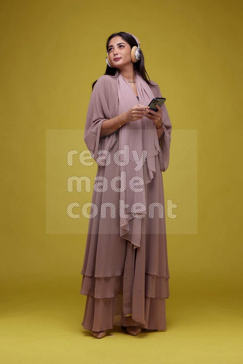 A Saudi woman Listing to Music on a Yellow Background wearing Brown Abaya