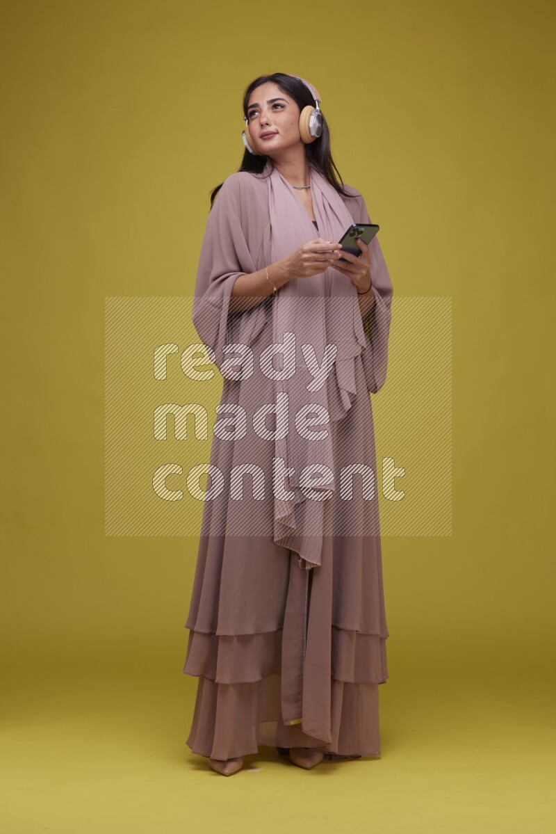 A Saudi woman Listing to Music on a Yellow Background wearing Brown Abaya