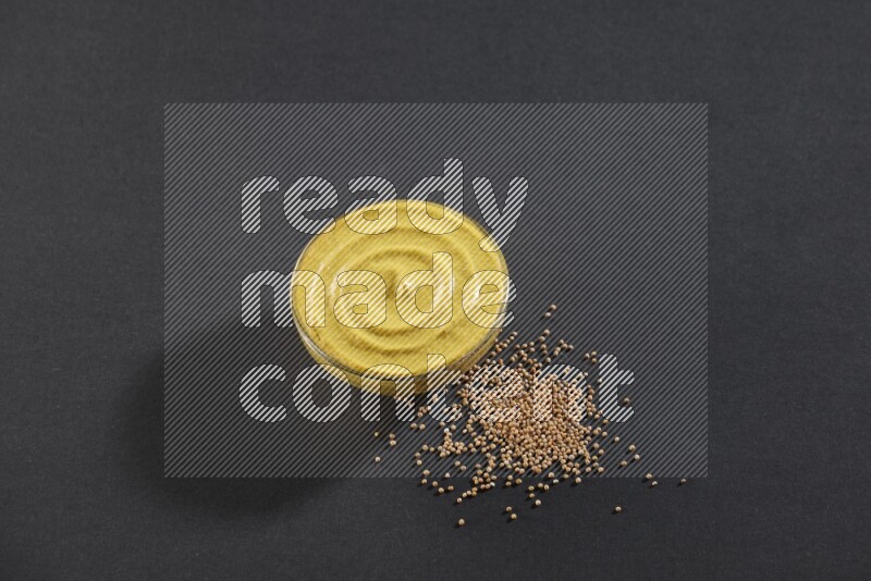 A glass bowl full of mustard paste with mustard seeds underneath on black flooring