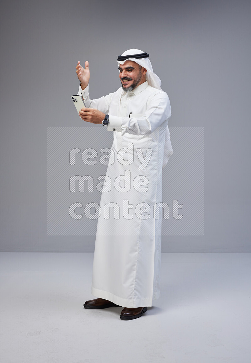 Saudi man Wearing Thob and white Shomag standing texting on phone on Gray background