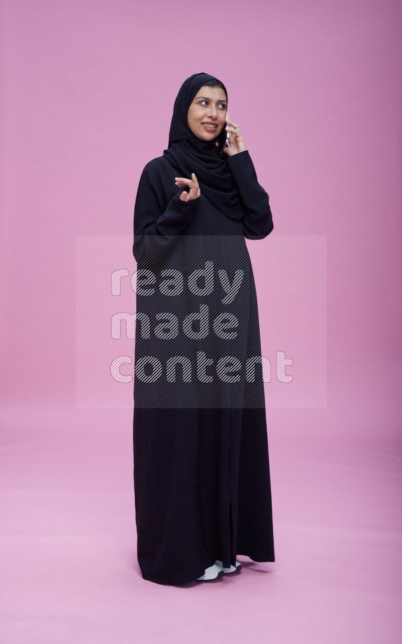 Saudi woman wearing Abaya standing talking on phone on pink background
