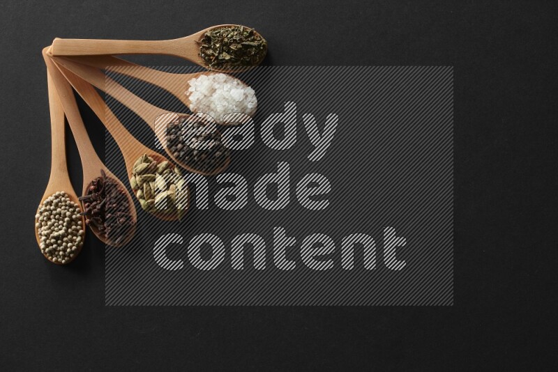 الفلفل الأبيض والقرنفل والهيل والملح والفلفل الأسود وريحان مجفف في معالق خشبية علي خلفية سوداء