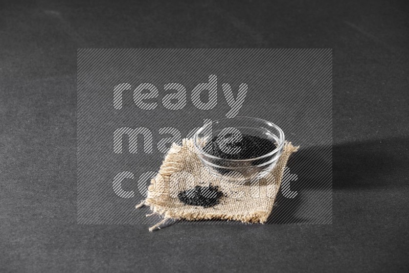 A glass bowl full of black seeds and seeds on burlap fabric on a black flooring