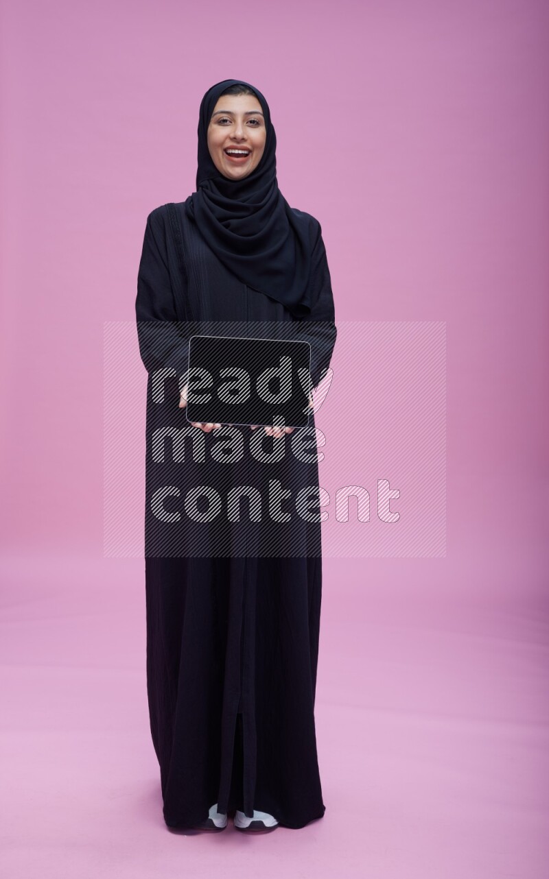 Saudi woman wearing Abaya standing showing tablet to camera on pink background