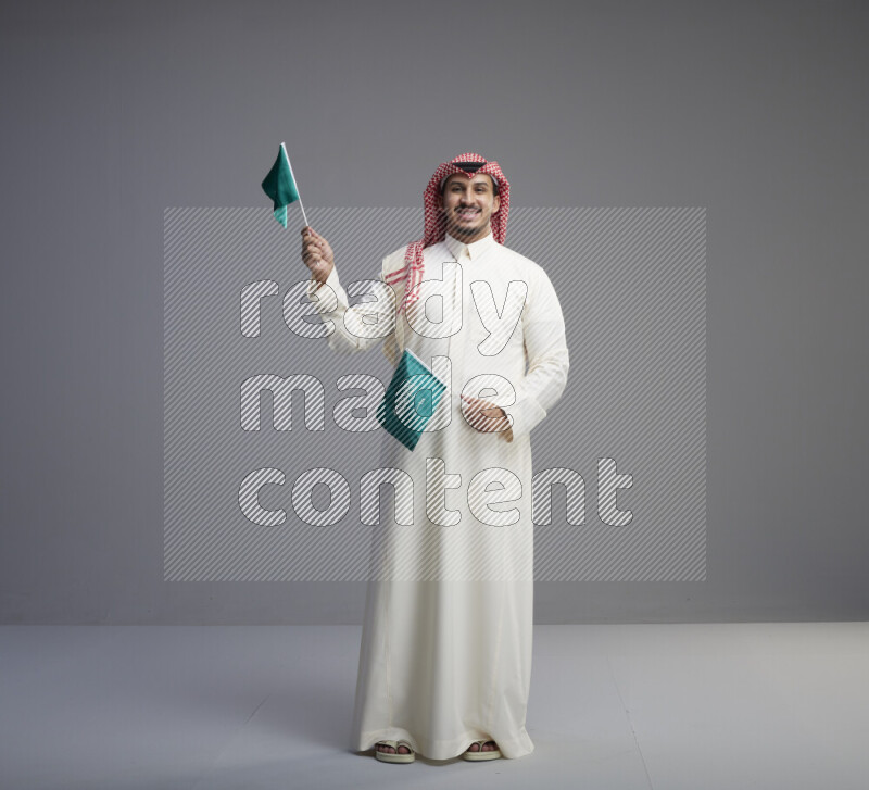 A Saudi man standing wearing thob and red shomag raising small Saudi flag on gray background