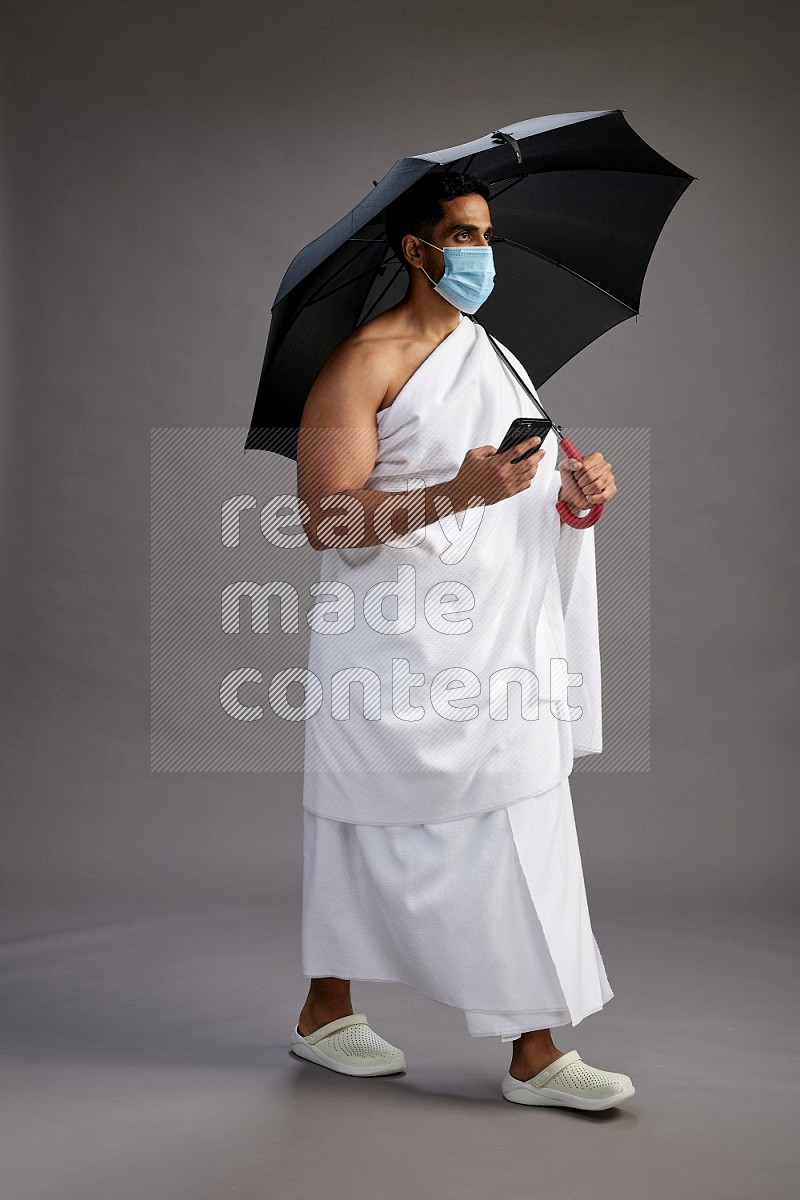 A man wearing Ehram and mask Standing holding umbrella on gray background on gray background