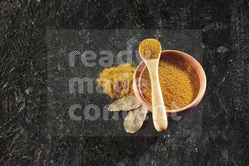 A wooden bowl and a wooden spoon full of turmeric powder on textured black flooring