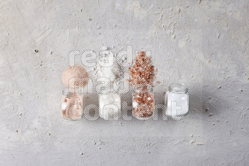 4 glass jars filled with table salt, coarse sea salt, fine himalayan salt and coarse himalayan salt on white background