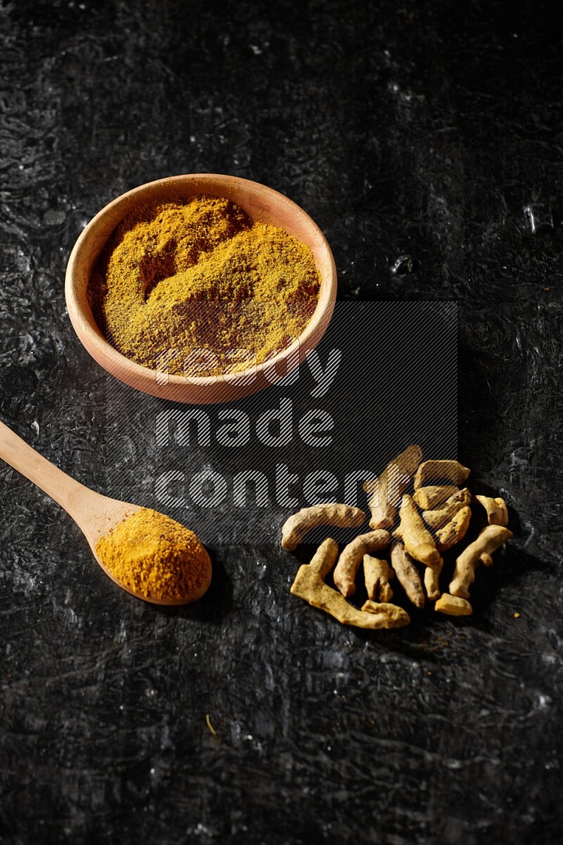 A wooden bowl and a wooden spoon full of turmeric powder with dried turmeric fingers on textured black flooring