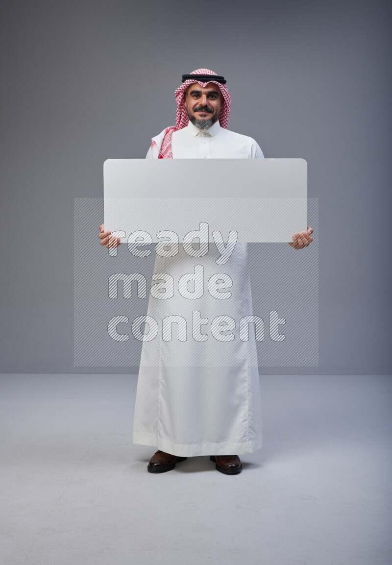Saudi man Wearing Thob and red Shomag standing holding board on Gray background