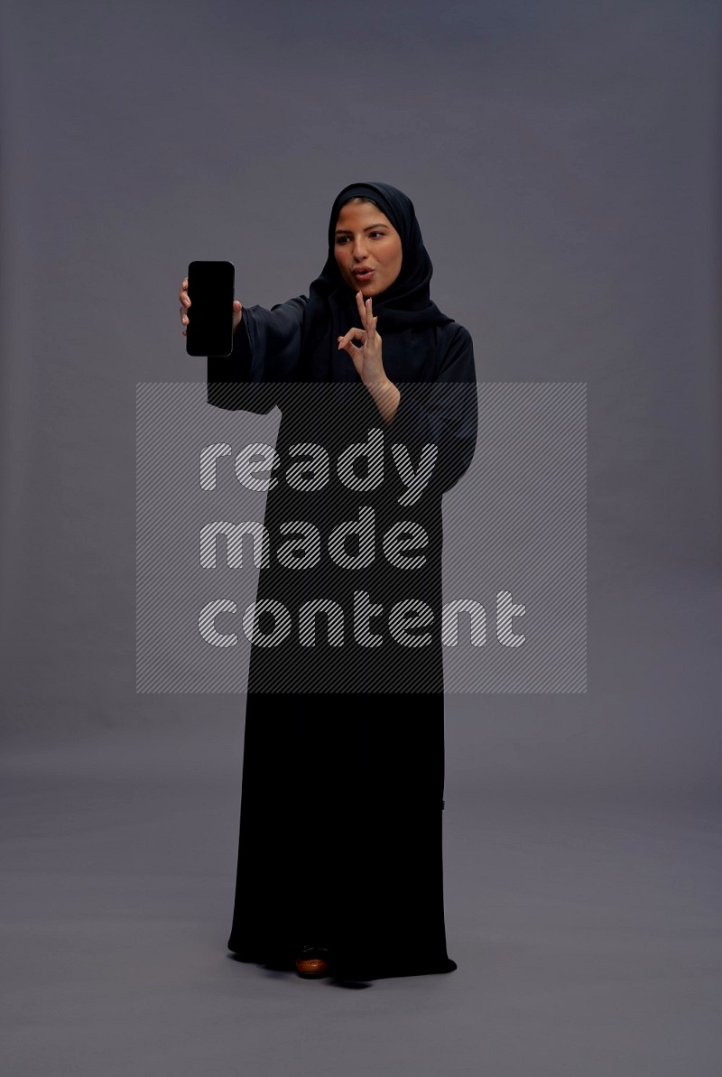 Saudi woman wearing Abaya standing showing phone to camera on gray background