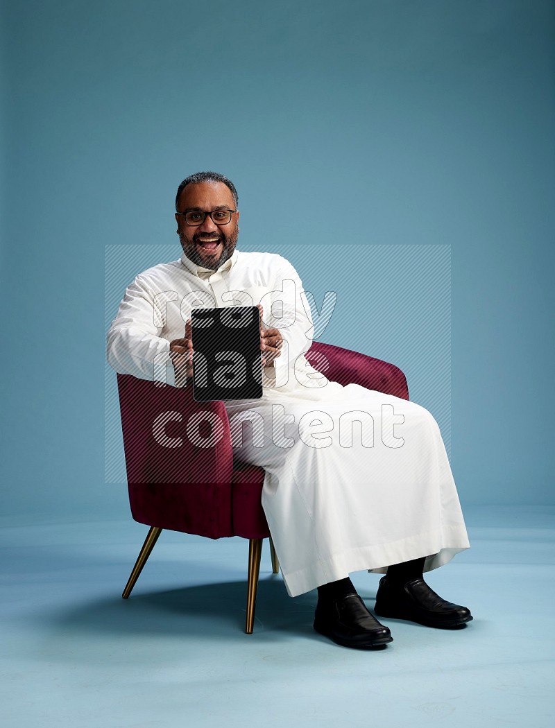 Saudi Man without shimag sitting on chair working on tablet on blue background
