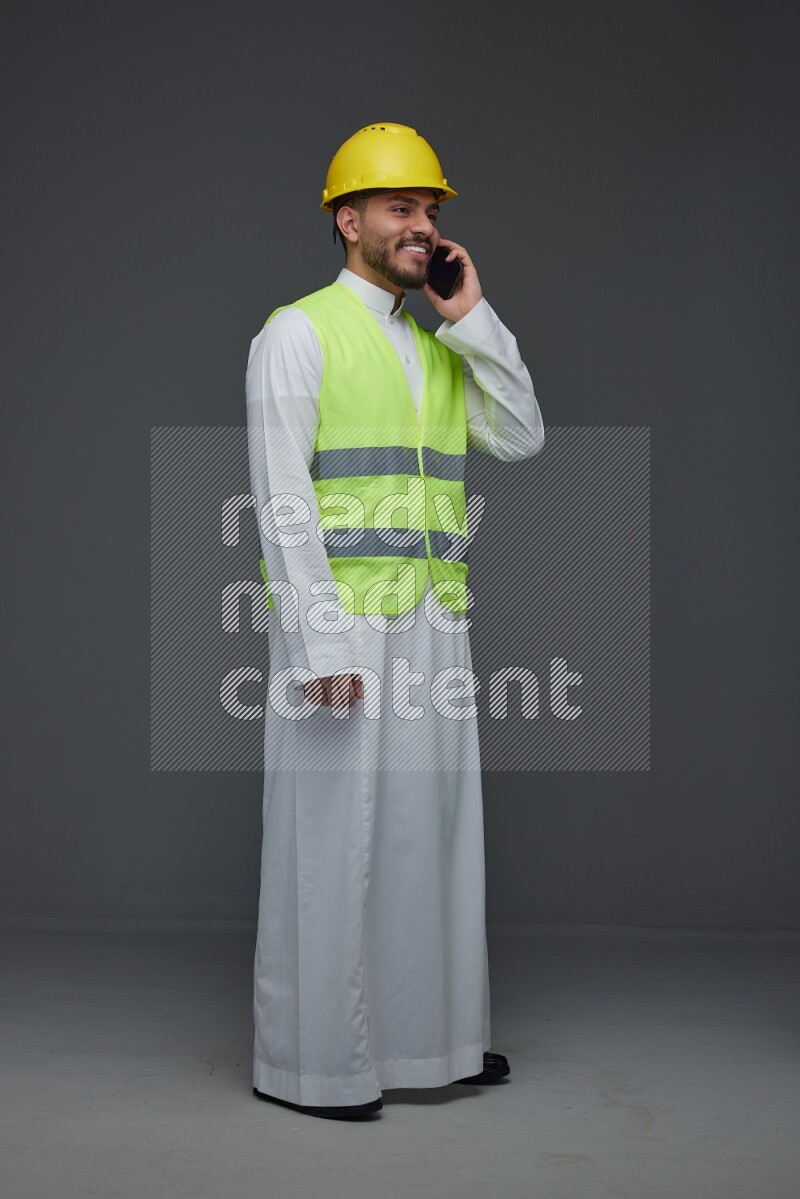 A Saudi man wearing Thobe with a yellow safety vest and white helmet standing and talking in the phone eye level on a gray background