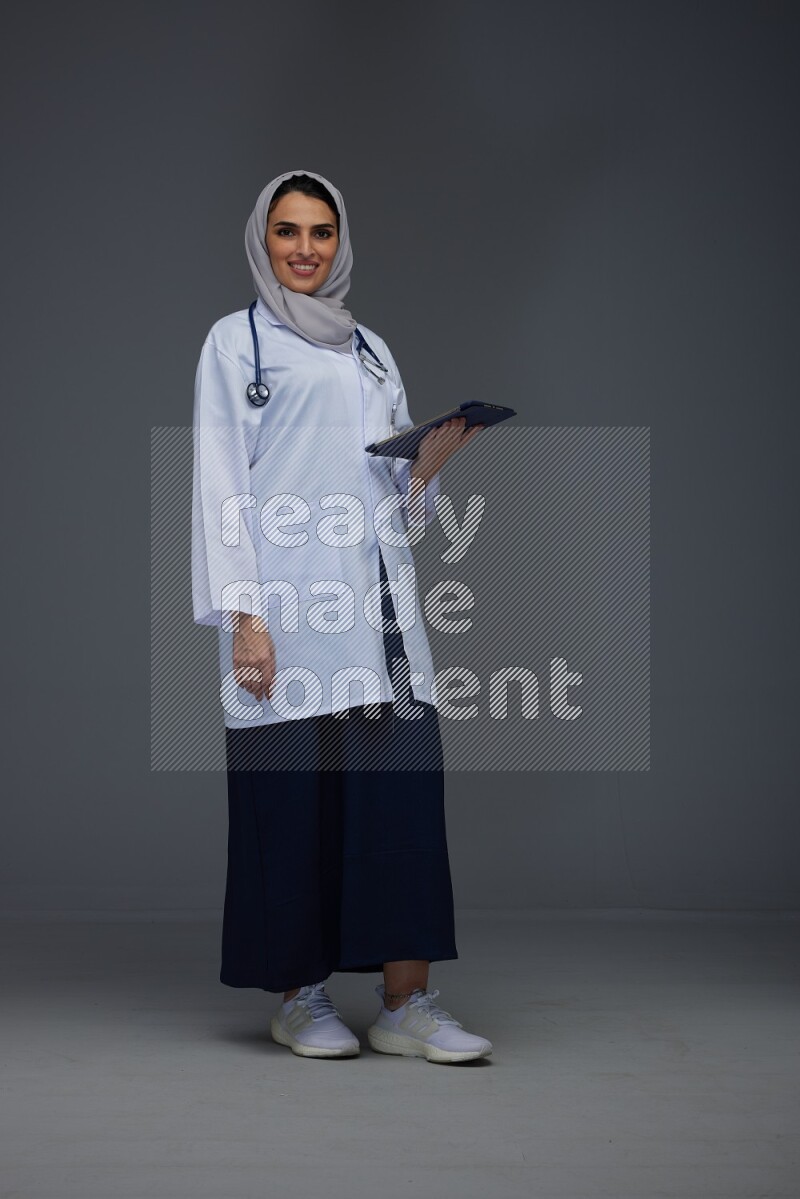 A Saudi doctor wearing a light gray head scarf standing and crossing her hands eye level on a grey background