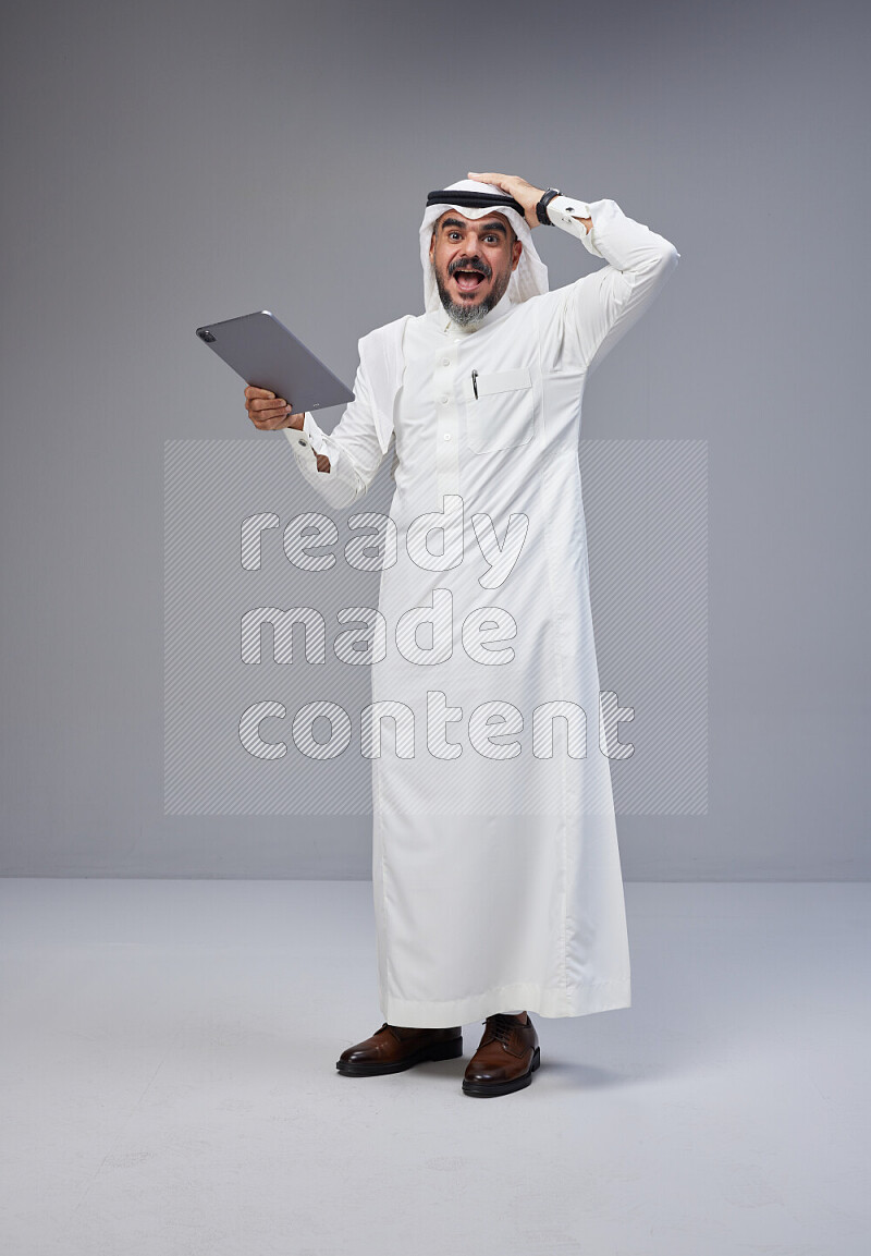 Saudi man Wearing Thob and white Shomag standing working on tablet on Gray background