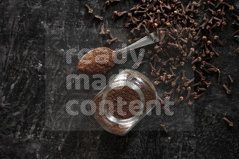 A glass spice jar and a metal spoon full of cloves powder with cloves spread on textured black flooring