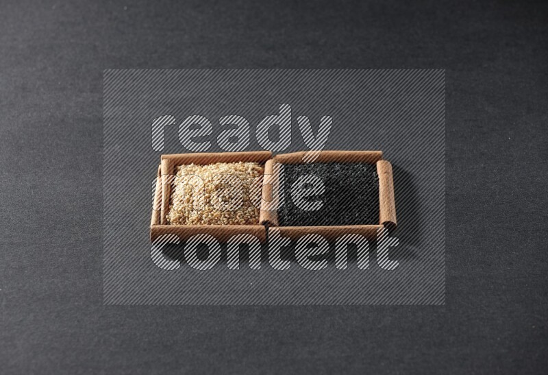 مربعان من أعواد القرفة ممتلئان ببذور السمسم وحبة البركة السوداء علي خلفية سوداء