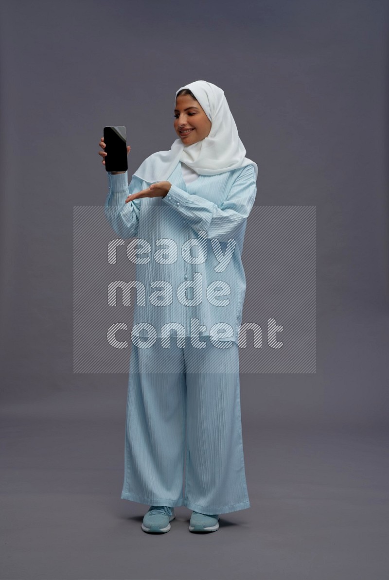 Saudi woman wearing hijab clothes standing showing phone to camera on gray background
