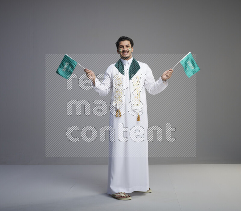 A Saudi man standing wearing thob and saudi flag scarf with face painting holding small Saudi flag on gray background