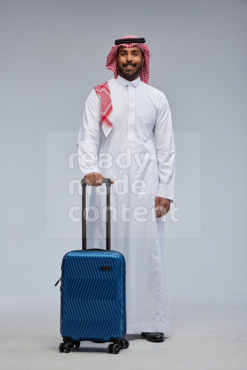 Saudi man wearing Thob and shomag standing holding Travel bag on Gray background