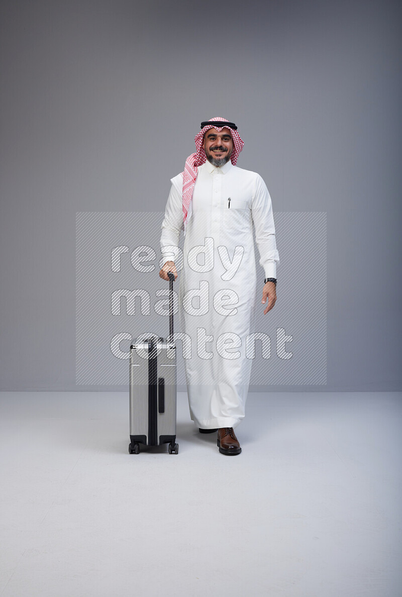Saudi man wearing Thob and red Shomag standing holding Travel bag on Gray background