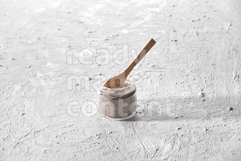 A glass jar full of black pepper powder with a wooden spoon on a textured white flooring
