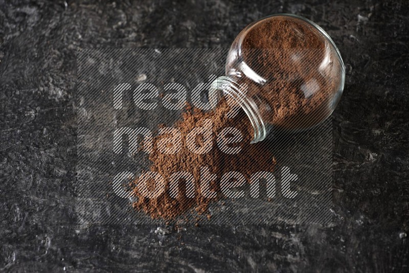 A flipped glass spice jar full of cloves powder and powder came out of it on textured black flooring