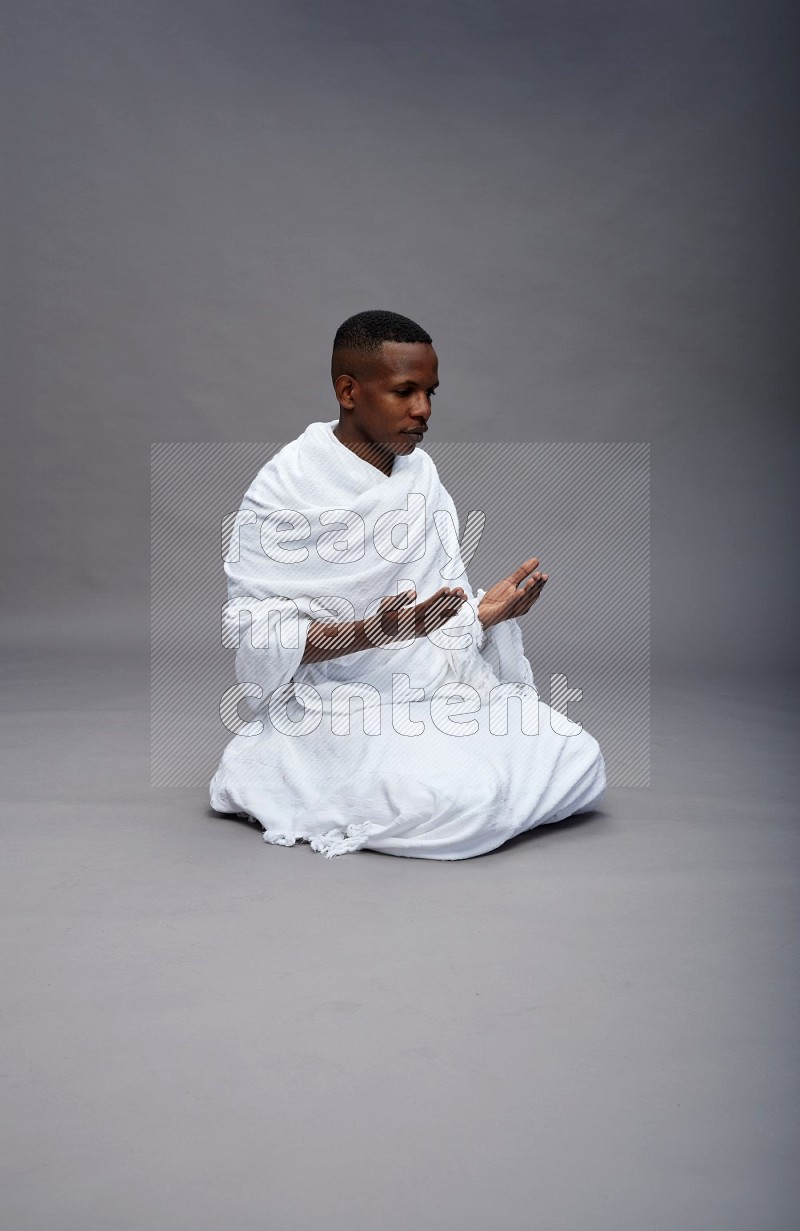 A man wearing Ehram sitting on floor dua'a on gray background