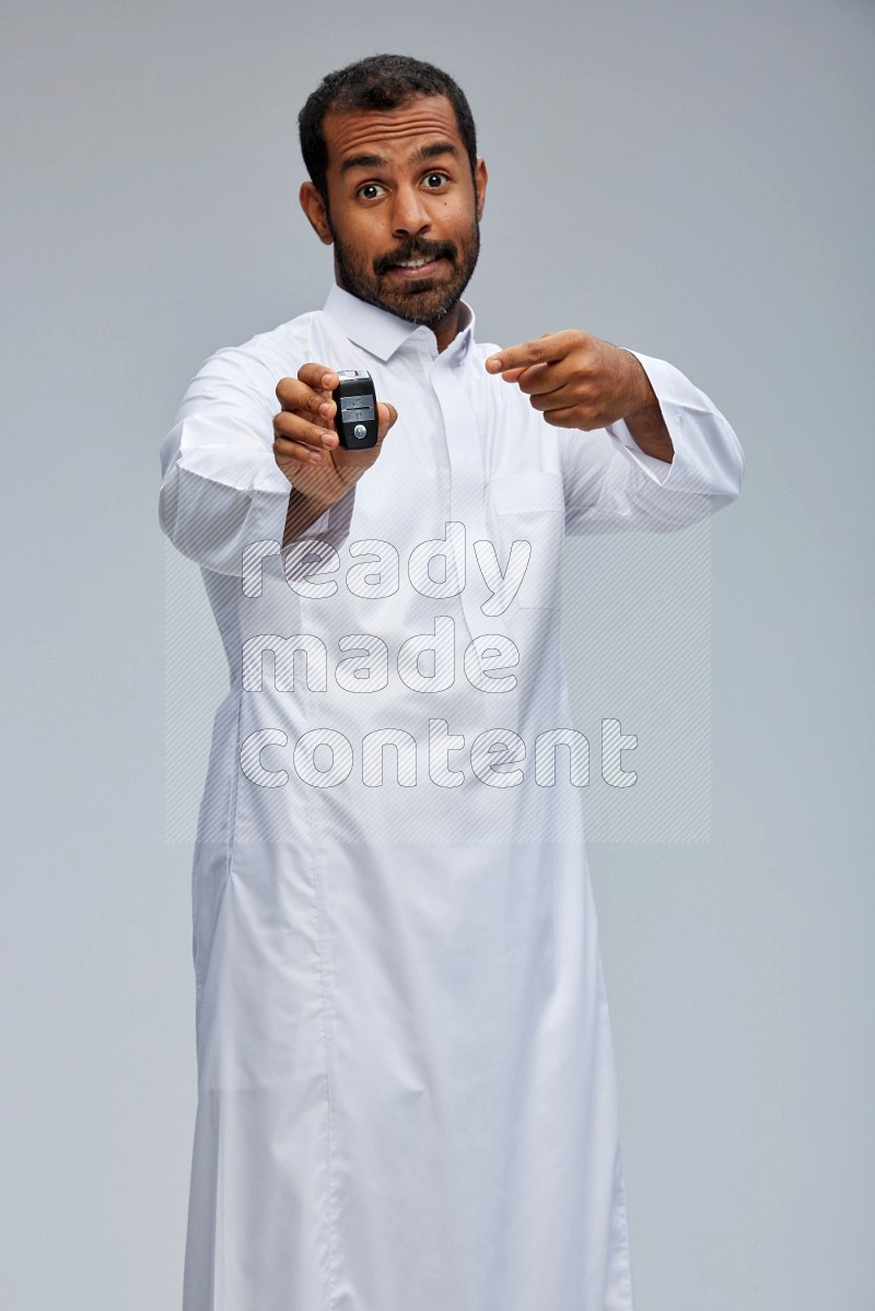Saudi man wearing thob and shomag standing holding car key on gray background