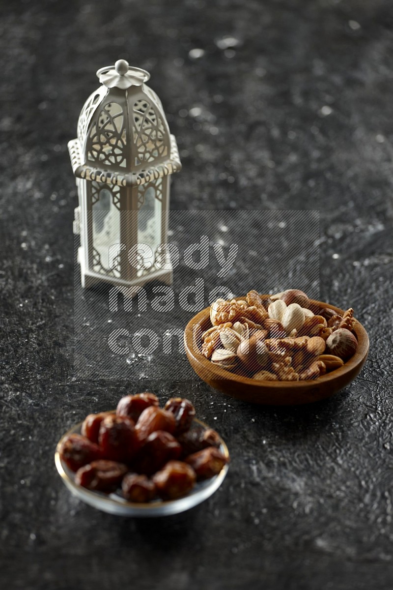 A white lantern with different drinks, dates, nuts, prayer beads and quran on textured black background