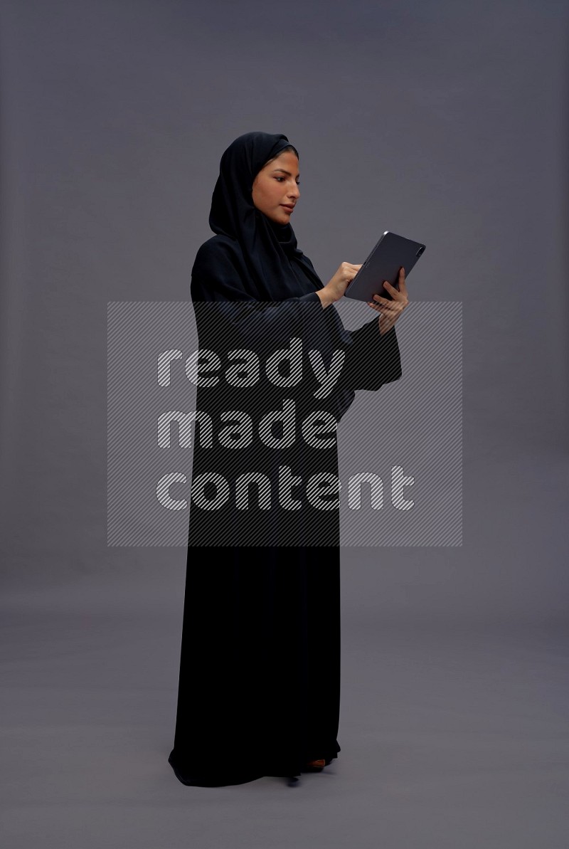 Saudi woman wearing Abaya standing working on tablet on gray background