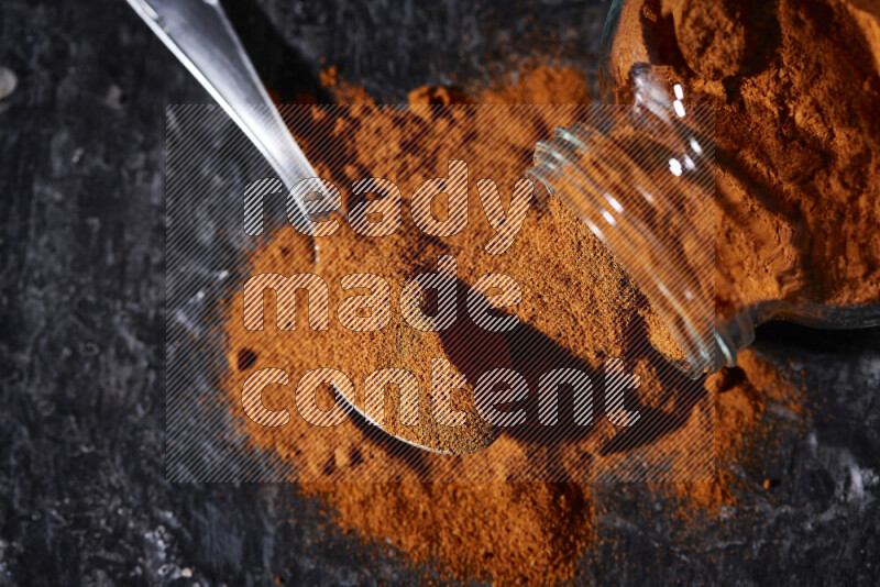 A glass jar full of ground paprika powder flipped with some spilling powder on black background