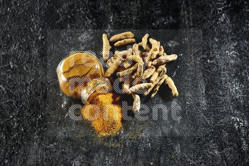 A flipped glass spice jar full of turmeric powder and powder spilled out of it with dried whole fingers on textured black flooring